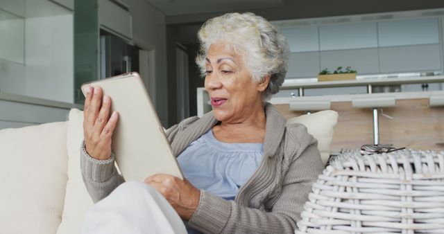 Elderly woman comfortably seated in a living room using a digital tablet, appearing engaged and smiling. This image can be used for depicting senior citizens embracing modern technology, connectivity, digital engagement, and leisurely activities at home.