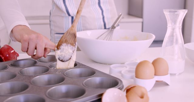 A person wearing white apron is scooping batter into a greased muffin tray using a wooden spoon. Surrounding the person are baking ingredients and utensils including flour, eggs, and various mixing bowls. This is ideal for content representing home baking, cooking tutorials, or culinary blog visuals.