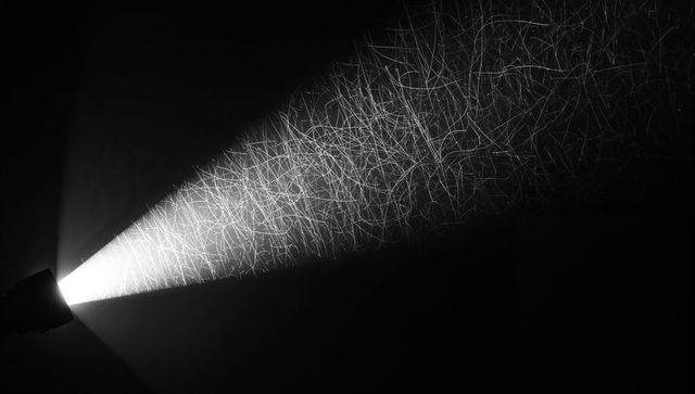 Handheld flashlight casting triangular beam revealing dust particles and fibers