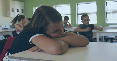 Pensive student resting head on desk while classmates using phones in bright classroom