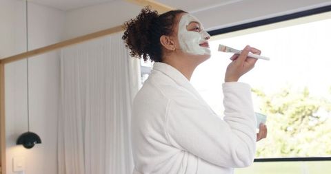 African american woman applying face mask in white bathrobe by sunlit window for self-care