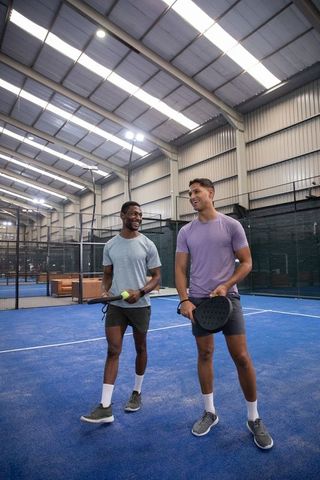 Diverse male friends walking and chatting on indoor padel court holding paddles