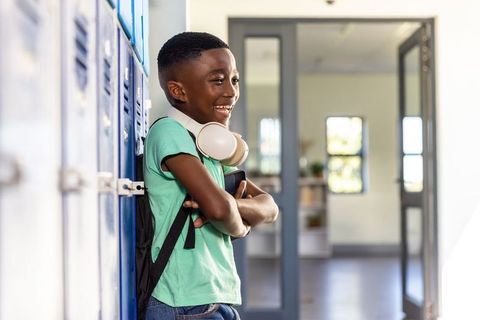 Smiling african american schoolboy leaning on lockers wearing headphones holding tablet