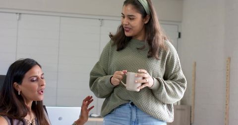 Multicultural female coworkers chatting in casual office holding mug and laptop