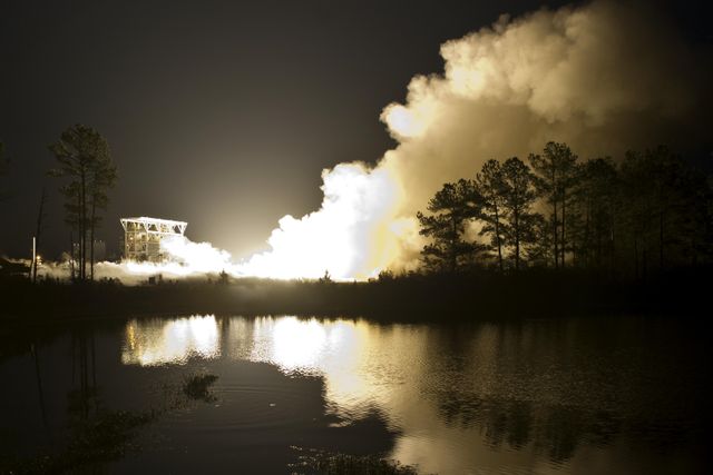 This image captures the impressive nighttime test fire of the Aerojet AJ26 engine at Stennis Space Center, with reflections on the adjacent lake and a smoke column. Ideal for use in articles about space exploration, rocket engineering, commercial spaceflight partnerships, and advancements in aerospace technology.