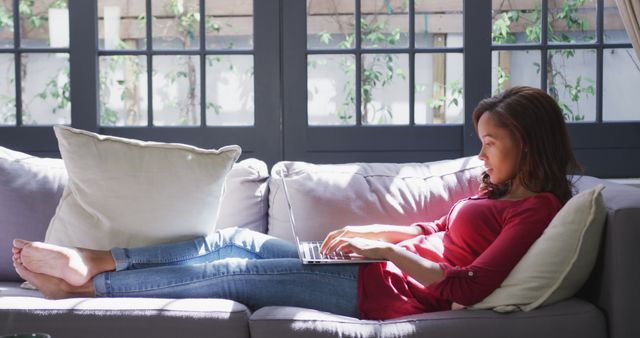 Woman lounging on light grey sofa, working on laptop. Room is well-lit from large windows, creating a cozy and inviting atmosphere. Ideal for promoting home comfort, remote work setups, or casual lifestyle content.