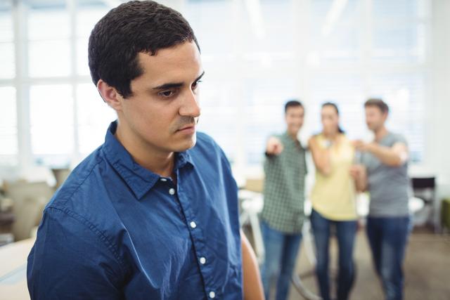 A man in a blue shirt is looking sad while three colleagues in the background are pointing and laughing at him. This image can be used to illustrate workplace bullying, harassment, and the emotional impact of unkind behavior in a professional environment. It is suitable for articles, blogs, and campaigns focused on promoting a healthy and respectful workplace culture.