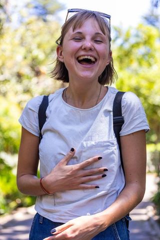 Young woman laughing and holding chest on sunlit park path, casual backpack