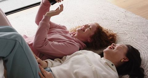 Diverse female friends laughing and sharing smartphone while lying on sunlit rug