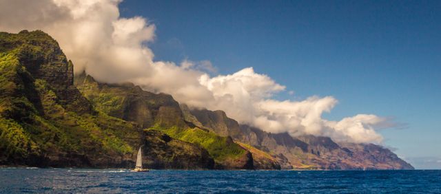 Sailboat gliding along clear blue ocean near Na Pali Coast on bright sunny day, with rugged cliffs reaching into clouds. Ideal for travel, adventure, and nature themes to evoke sense of exploration and scenic beauty.
