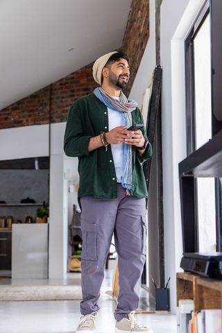 Freelancer standing by loft window holding smartphone, wearing beanie and scarf