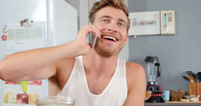 A man in a sleeveless shirt happily talking on the phone in a kitchen. Ideal for promoting home appliances, communication services, or lifestyle brands focused on casual, everyday moments. The bright and cheerful atmosphere is suitable for consumer goods advertising and social media content.