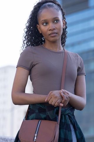 African woman standing in cityscape wearing brown top, plaid shirt tied, crossbody bag