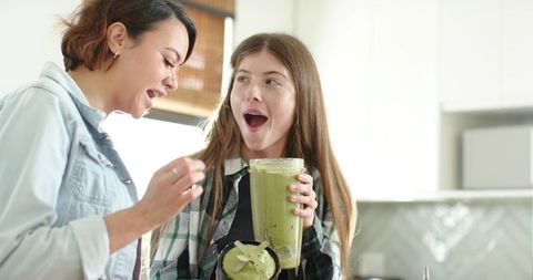 Diverse women tasting green smoothie in bright kitchen holding blender cup and spoon