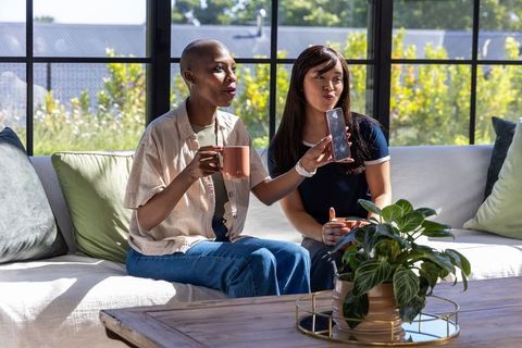 Sharing coffee and smartphone on sunlit sofa, diverse friends enjoying cozy modern home