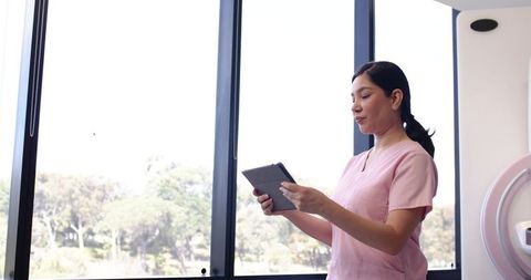 Female healthcare worker in pink scrubs using tablet beside radiology gantry by window