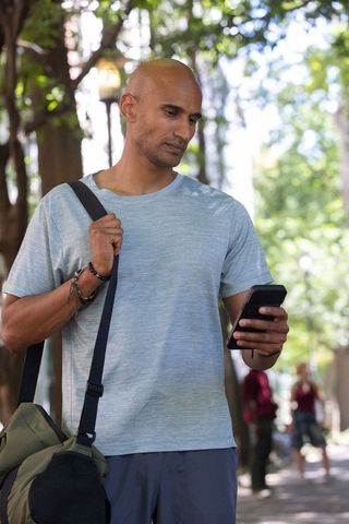 African man standing on shaded path using smartphone and carrying duffel bag, commute