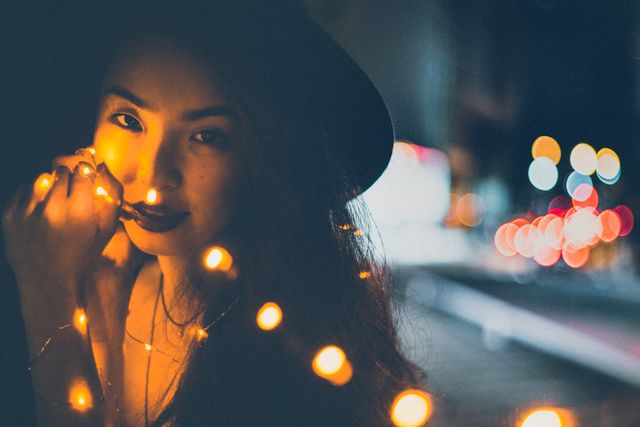 Woman in a hat adorned with glowing string lights, set against an urban nightscape with vibrant bokeh city lights. Perfect for use in creative, fashion, or urban lifestyle projects, showcasing modern nightlife or whimsical moments.
