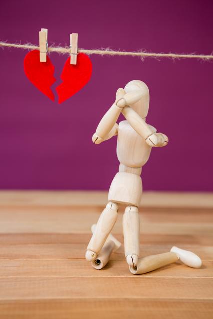 This image depicts a wooden figurine kneeling in front of a broken red heart hanging on a string, symbolizing heartbreak and emotional distress. The dramatic pose and vibrant colors make it suitable for illustrating themes of love, relationships, and emotional struggles. Ideal for use in articles, blog posts, and social media content related to romance, breakups, and mental health.