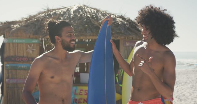 Two happy biracial men standing with surfboard on beach and talking. Vacations, holidays, spending time with friends.