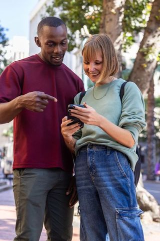 Smiling young couple sharing smartphone on sunny urban sidewalk, casual street style