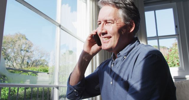 Mature man talking on phone while smiling, sitting by bright window with natural light. Useful for lifestyle, communication, home living, and technology-related content.