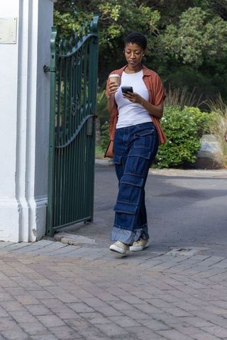 African american woman walking through garden gate, checking phone while holding coffee