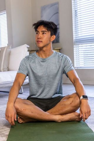 Asian man sitting cross-legged on mat practicing mindful yoga at home wearing smartwatch