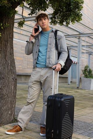 Late-teen traveler talking on smartphone while holding carry-on suitcase and backpack