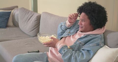 Mid-adult african american man relaxing on sofa with popcorn watching tv in pink hoodie