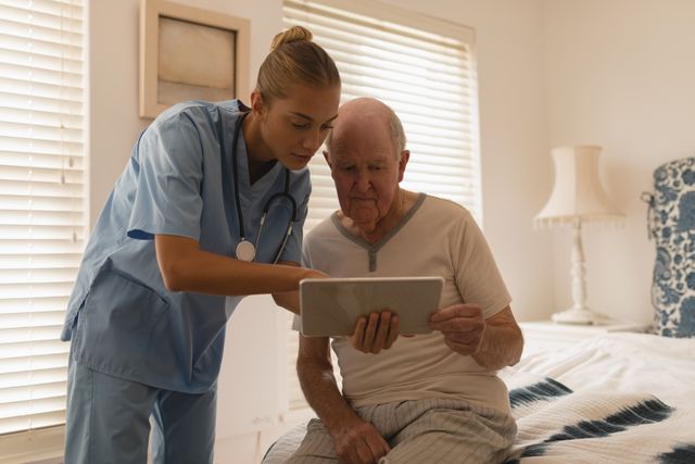 Front view of a female doctor using digital tablet and showing something to a senior man sitting on bedroom at home