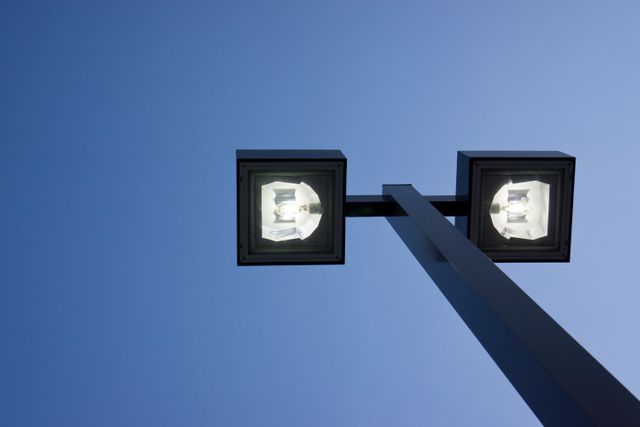 Double-headed street lamp shining brightly against a clear blue evening sky. This vibrant image of urban infrastructure may be used for topics related to city life, outdoor lighting, or technological advancements in public illumination.