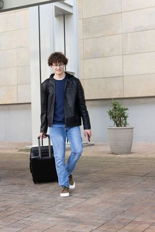 Young traveler walking through city plaza pulling wheeled suitcase holding smartphone