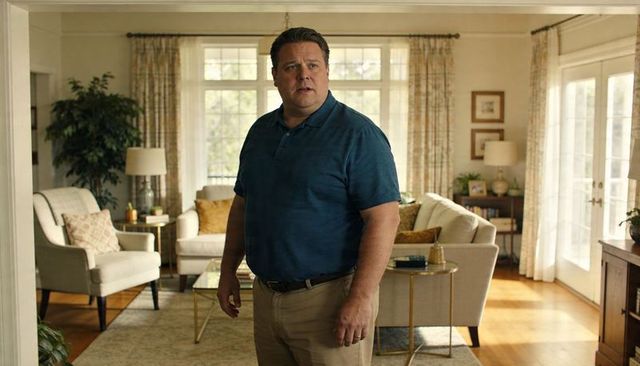Man standing in cozy sunlit living room wearing teal polo and khaki pants looking left