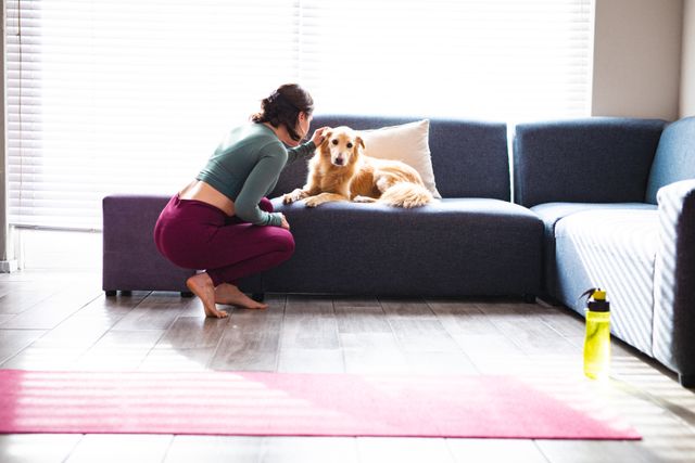 Caucasian woman taking a break from exercising to scratch her dog in a cozy living room. Ideal for use in articles or advertisements about home workouts, pet ownership, domestic life, and relaxation. Can also be used in content promoting healthy lifestyles, pet care, and home comfort.