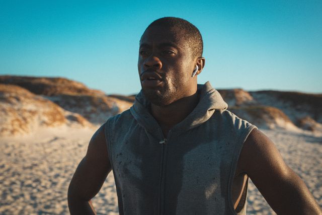 African American man resting after a run on the beach at sunset. He is wearing a sleeveless hoodie and earphones, with sweat visible on his face. This image can be used for promoting fitness, healthy lifestyle, outdoor activities, and exercise routines. Ideal for fitness blogs, health magazines, and advertisements for sportswear or fitness equipment.