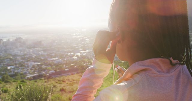 Woman adjusting earphones while standing on a hilltop with a scenic view of the city at sunrise. Ideal for illustrating fitness, morning routines, urban exercising, outdoor activities, motivational content, and lifestyle promotions.