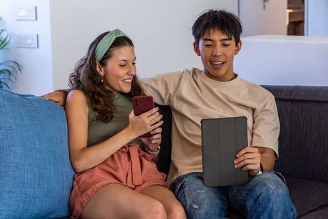 Diverse young couple sharing smartphone and tablet on couch, smiling and enjoying digital connection
