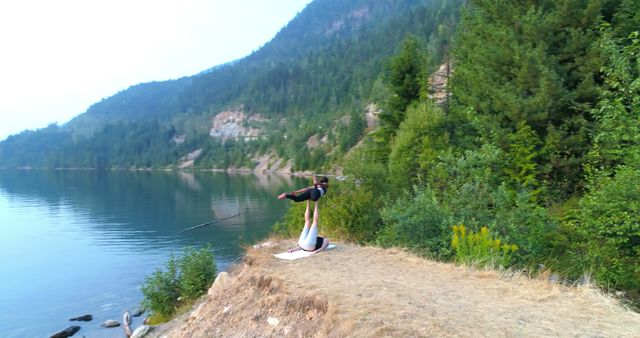 A couple practicing acroyoga on a cliff edge near a serene lake with lush green mountains and trees in the background. Ideal for themes related to outdoor activities, fitness, relationship bonding, and mindfulness retreats.