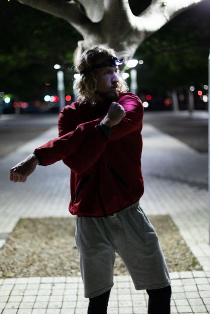 Fit man with long blonde hair stretching his arms outdoors in the city at night. He is wearing sportswear and a headlight, indicating he is preparing for or cooling down from a workout. Ideal for use in articles or advertisements related to fitness, nighttime exercise routines, urban workouts, or active lifestyle promotions.