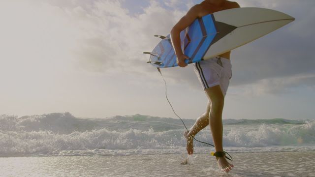 Shirtless male surfer running along sandy beach with surfboard, waves crashing in background, capturing energetic and adventurous mood of surfing lifestyle. Ideal for use in travel brochures, beach activity promotions, or inspirational content showcasing freedom and excitement.