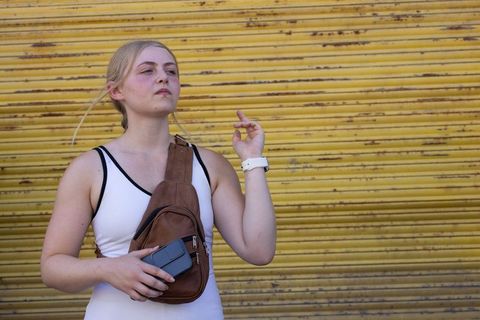 Young woman standing by yellow shutter wearing white tank and sling bag holding phone