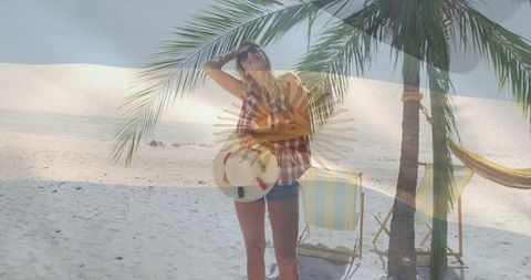 Standing woman wearing sunglasses holding hat and swim ring under palm with sun flag overlay