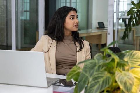 Indian businesswoman working at modern glass office desk with laptop and plants