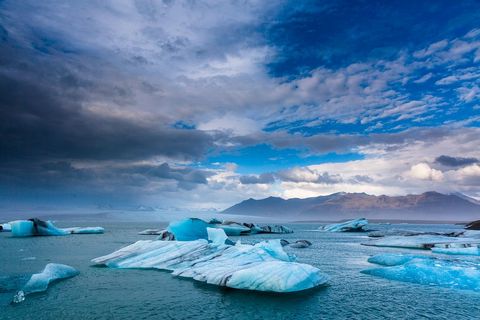 Blue Icebergs Floating in Glacial Lagoon under Dramatic Cloudscape with Distant Mountains