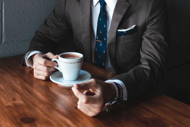 This image depicts a professional businessman in a suit holding a cup of coffee at a wooden desk. The setting looks like an office environment, conveying a sense of professionalism, routine, and break time at work. This can be used in business-related content, corporate training materials, articles related to professional life, and promotional materials for office-related products.