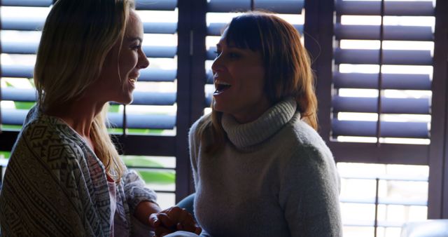 Two women are cheerfully chatting and laughing beside a window with blinds, creating a warm and friendly atmosphere. They are casually dressed in sweaters, indicating a relaxed indoor setting. This image can be used for topics related to friendship, bonding, casual conversations, and positive social interactions. It is also suitable for lifestyle blogs, promotional materials for social events, and articles about maintaining relationships.