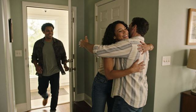 Warm homecoming hug between smiling couple while friend entering through open front door