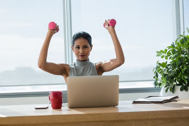 Portrait of smiling executive exercising with dumbbells while working on laptop in office