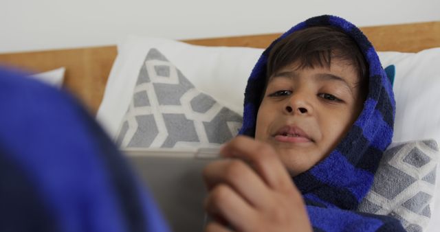 Boy in a blue bathrobe lies in bed and uses a tablet. Suitable for themes of technology use by children, indoor coziness, relaxation, and leisure activities. Ideal for family-oriented articles, tech reviews focusing on children's devices, and lifestyle blogs.